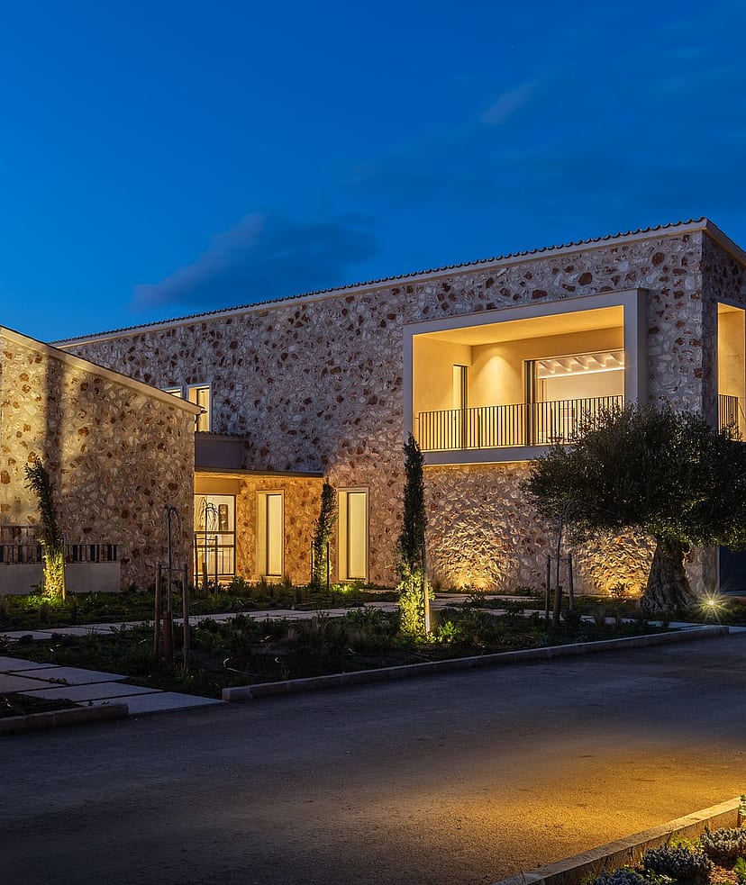 Exterior view of a modern stone-clad finca in Santanyí at twilight, showing illuminated interior spaces and landscaped grounds.