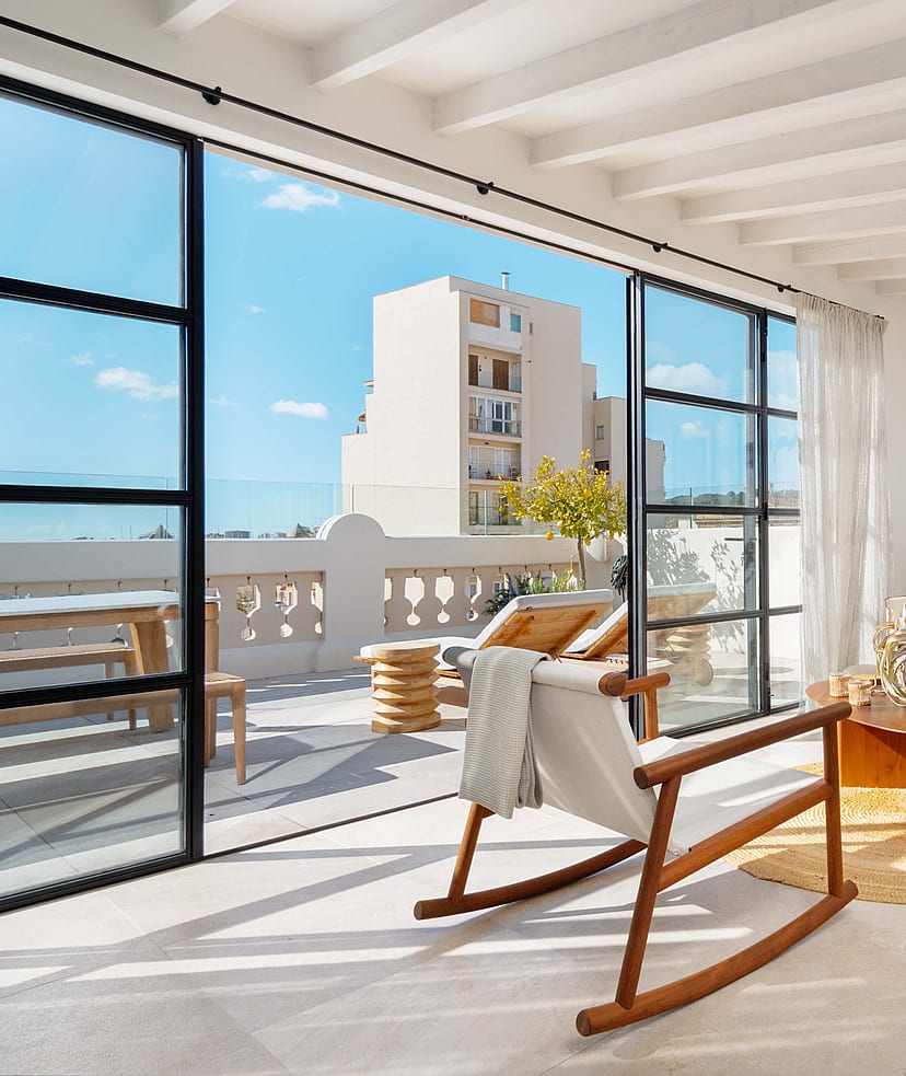 Upper-level sitting room with wooden rocking chairs and a cream sofa, steel-framed doors open to the terrace and rooftops beyond.