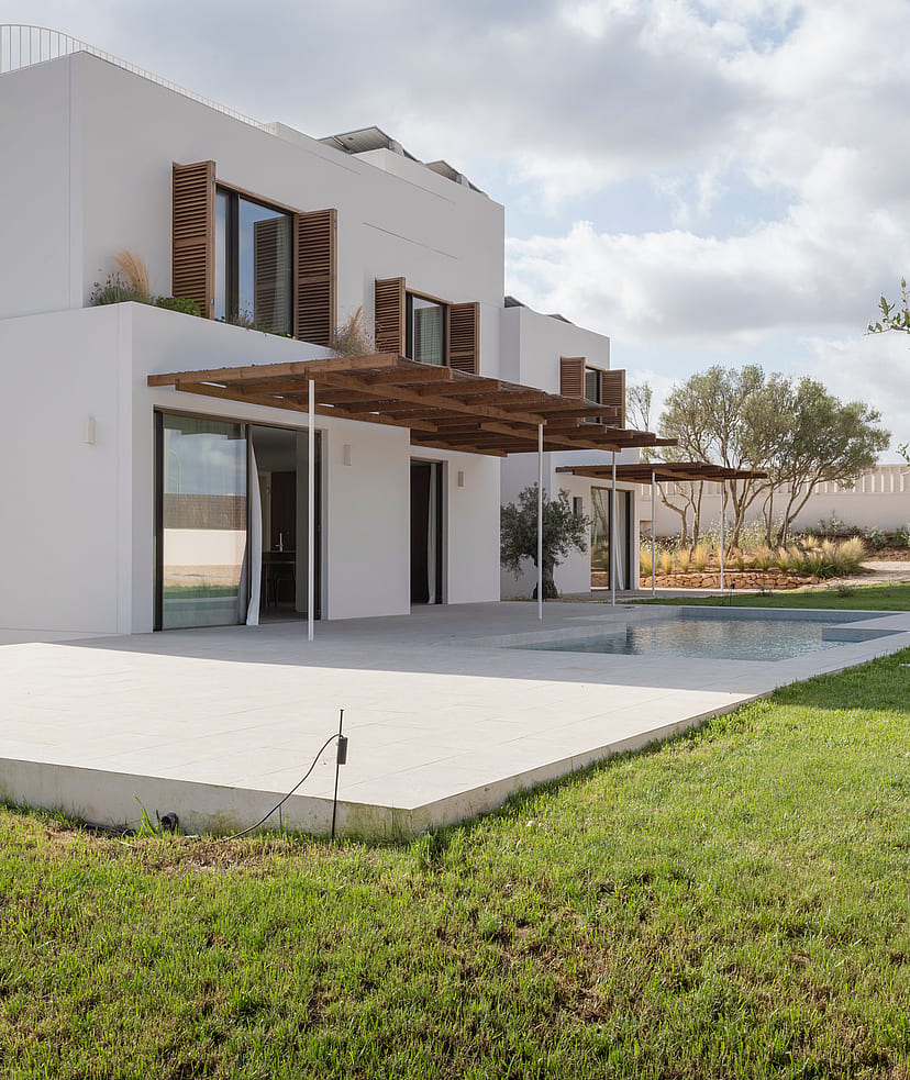 Modern two-story white villa with wooden shutters, a large pergola, and stone terrace overlooking a green lawn.
