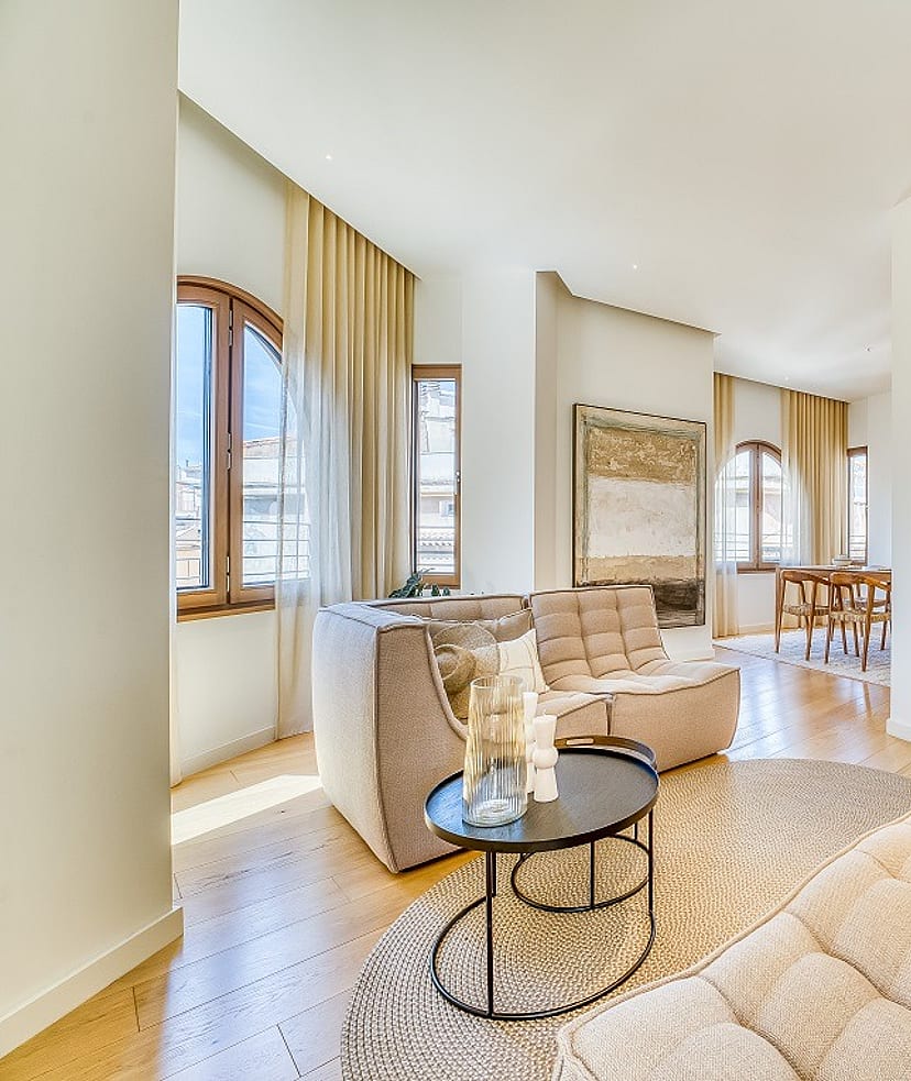 A bright living room in a penthouse with arched windows, light oak flooring, beige modular sofas, and a dining area in the background.