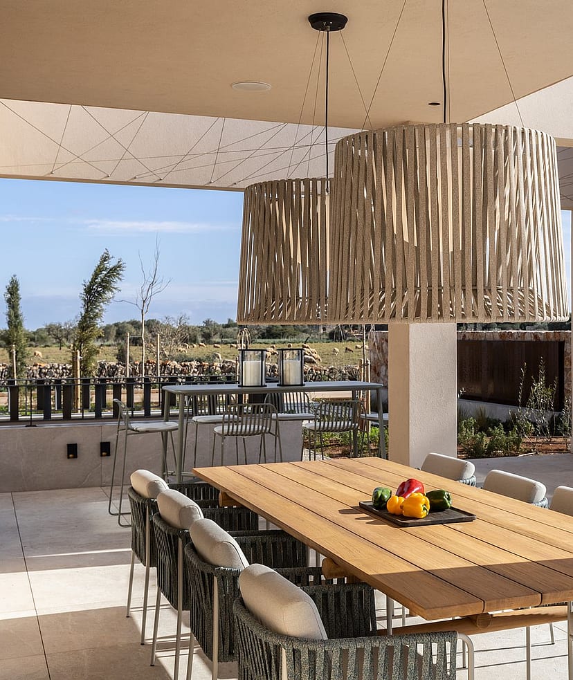 A spacious outdoor dining patio features a long wooden table, woven chairs, and a stone-built summer kitchen, looking towards mountain fields.