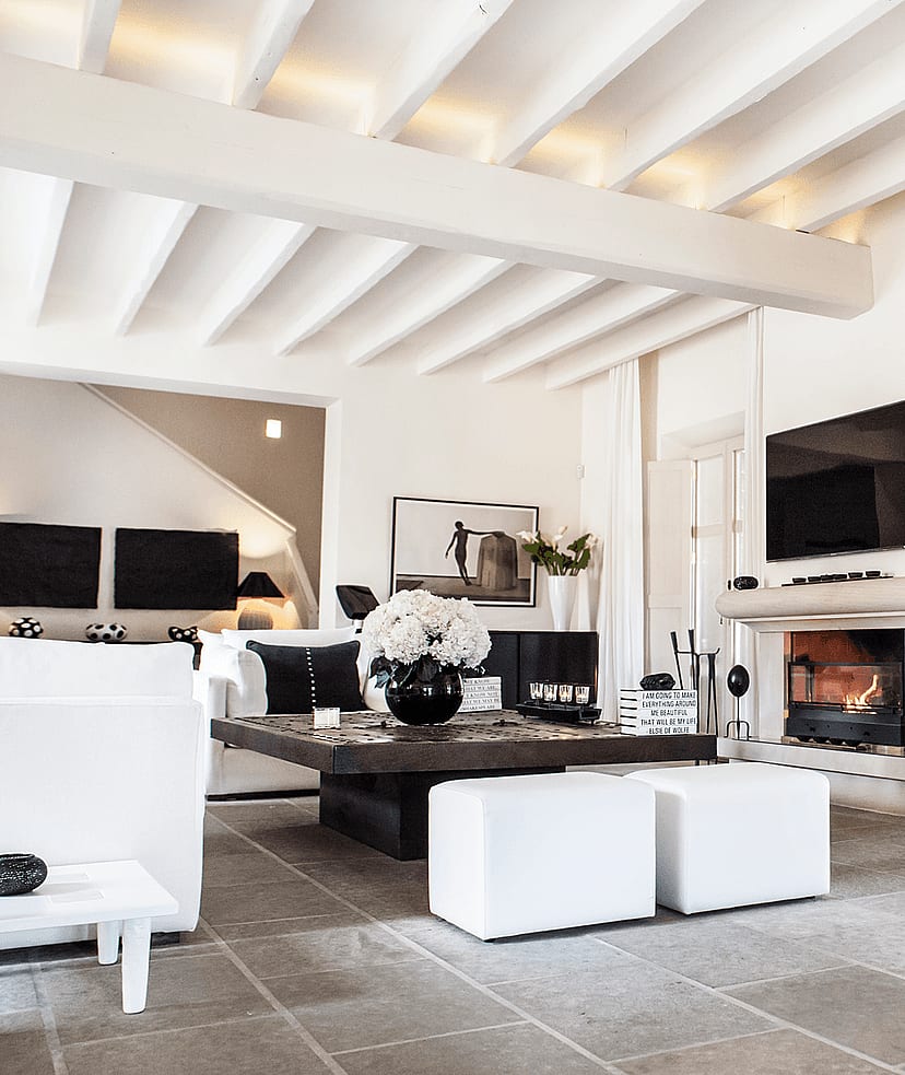 Bright living area with a stone fireplace, exposed beam ceiling, stone flooring, and white linen curtains.