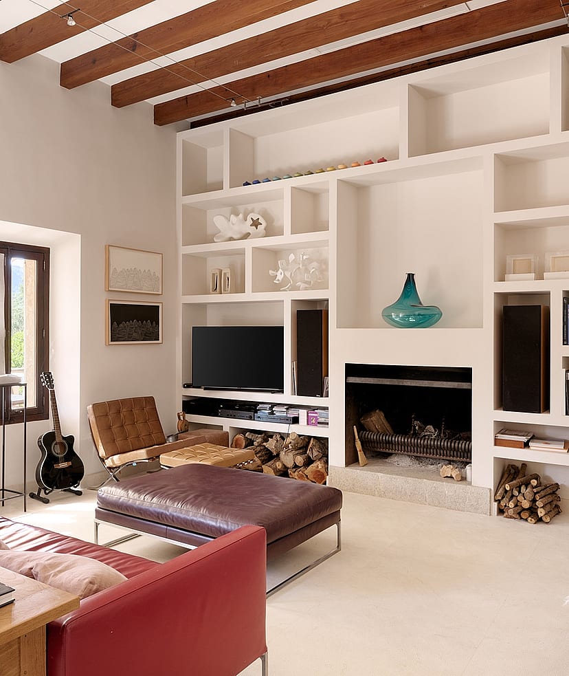 Living room with exposed wood ceiling beams, white built-in shelving unit, dark red sofa, and an open fireplace.
