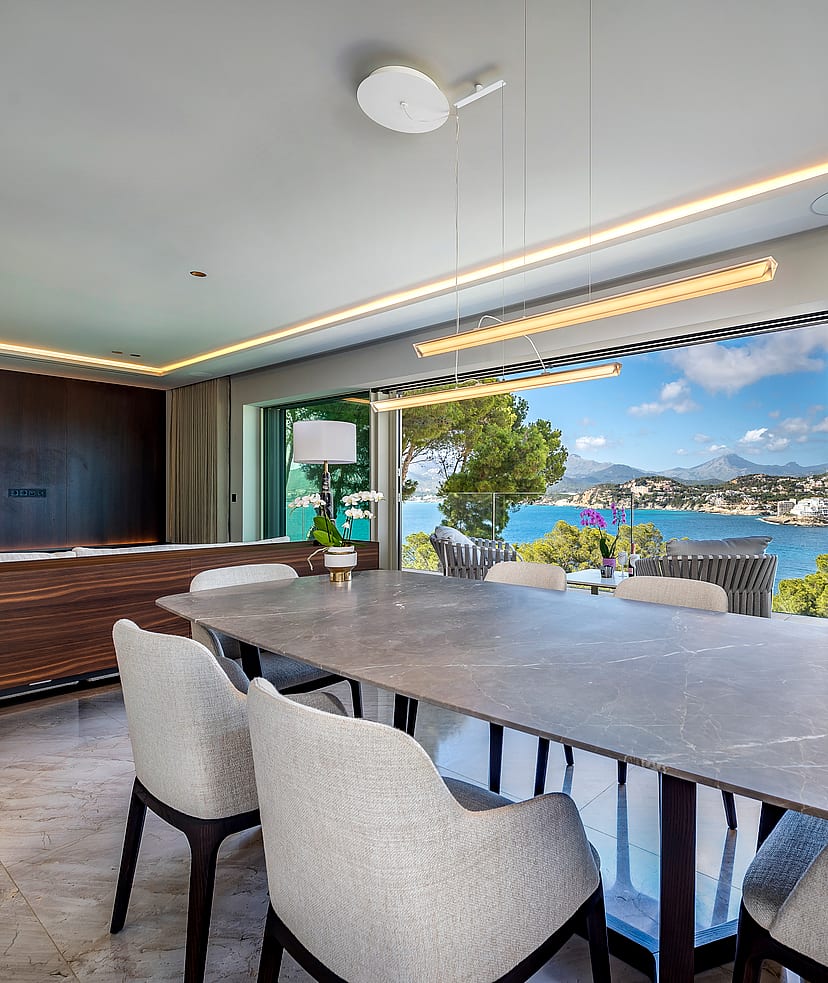 Dining room with a large oval grey stone table and upholstered chairs, facing an open terrace overlooking a blue bay.
