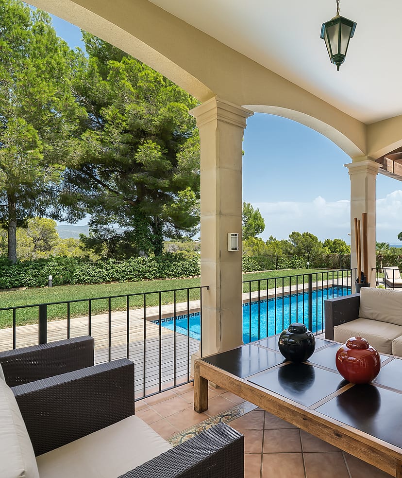 A covered terrace with comfortable outdoor seating overlooks a blue swimming pool and surrounding green gardens on a sunny day.