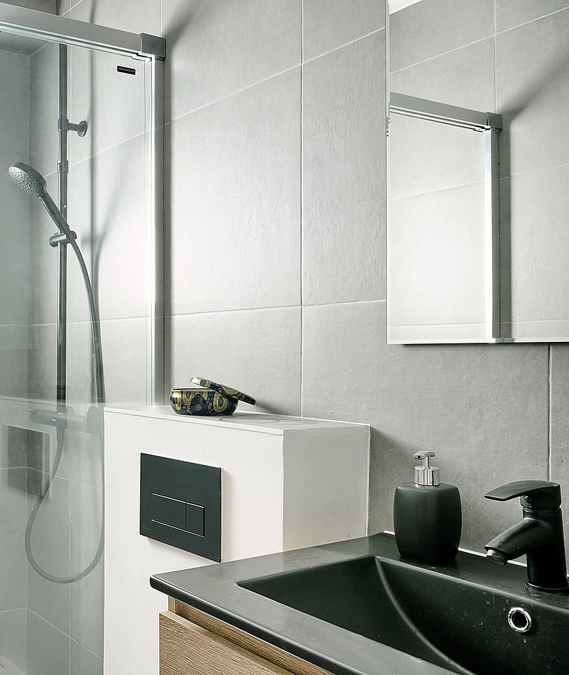 A bathroom with grey tiled walls, a glass-enclosed shower with a rainfall head, and a vanity with a black sink.
