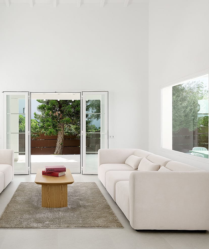 Salón blanco de doble altura con ventana arqueada, dos sofás color crema y puertas acristaladas hacia el jardín
