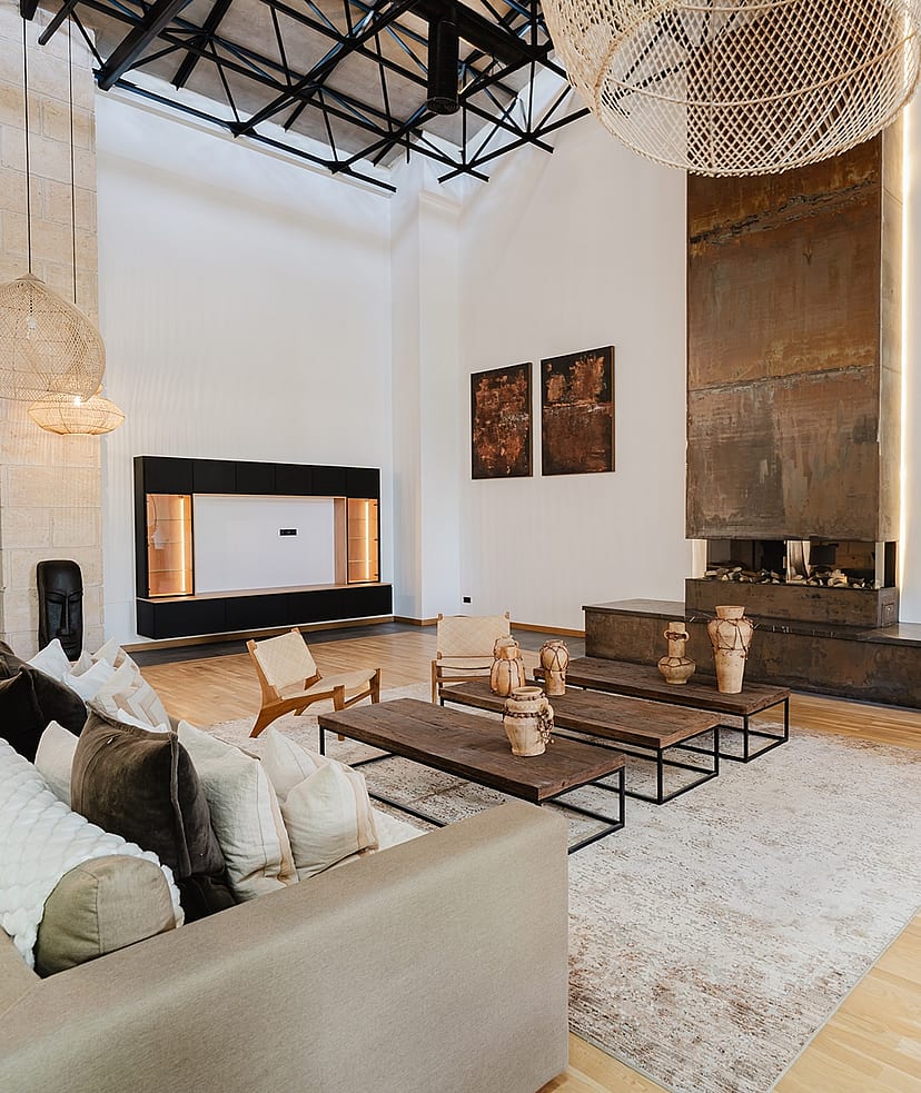 A spacious living area featuring a double-height ceiling with exposed black metal trusses, a tall rusted-metal fireplace surround, and woven pendant lighting.