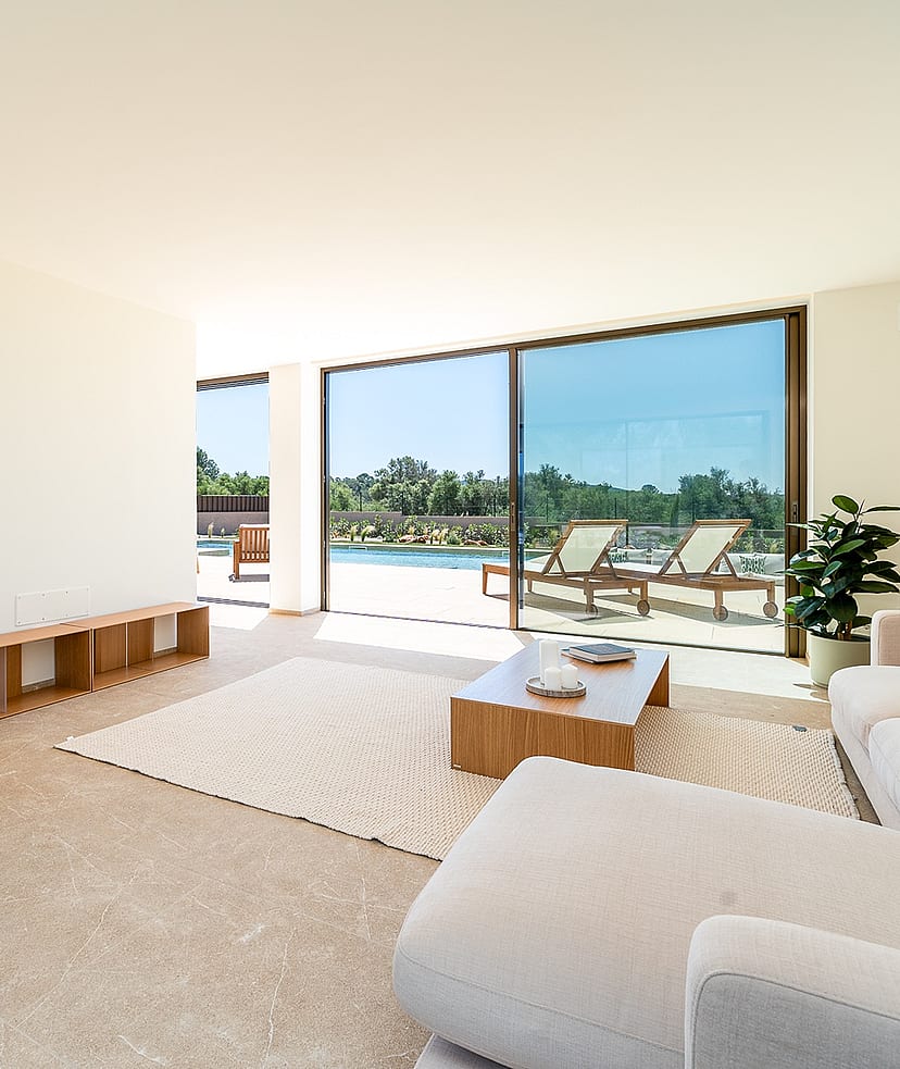 Bright open-plan living room featuring neutral sofas, a woven wood armchair, and floor-to-ceiling glass doors opening to a pool deck.
