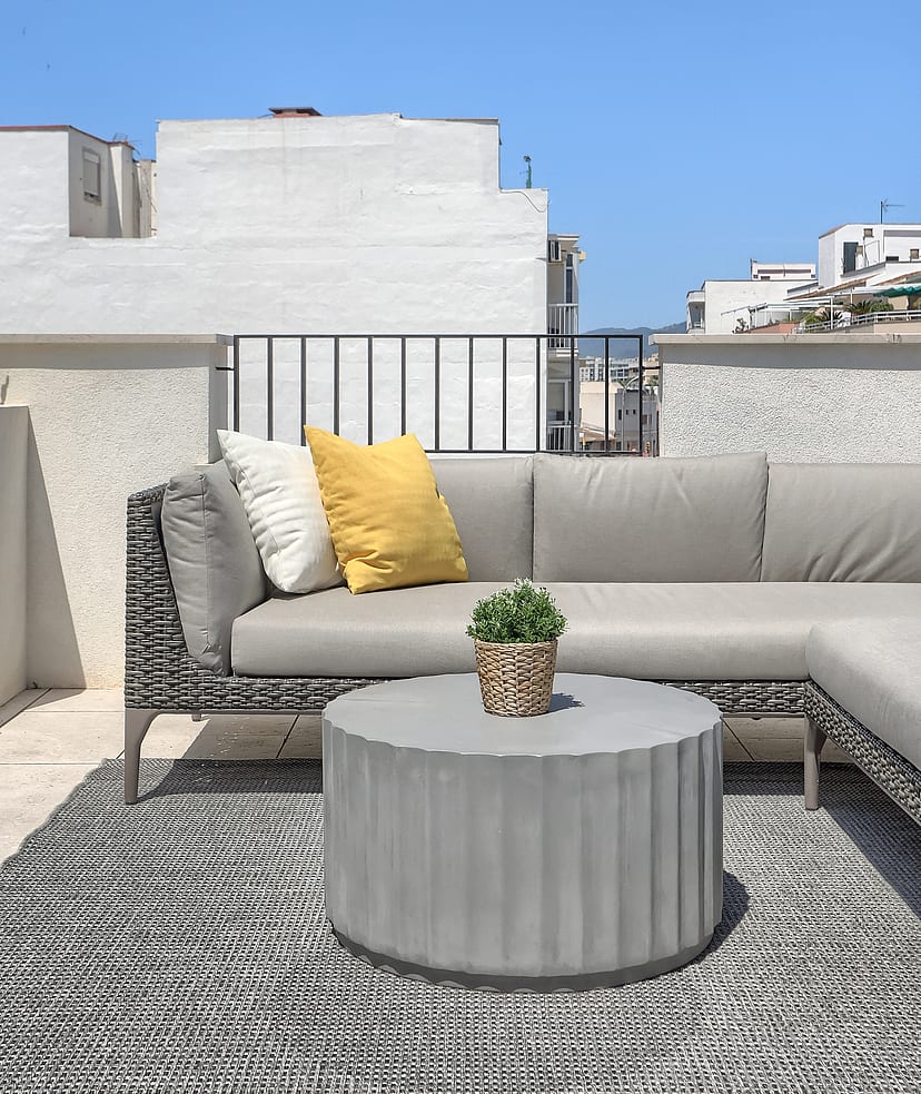 A rooftop terrace in El Molinar featuring a grey modular sofa with yellow cushions, a round fluted concrete table, and a patterned rug against a bright sky.