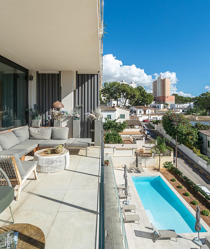 A covered terrace with grey sectional seating overlooks a rectangular swimming pool surrounded by a paved garden in a suburban setting.
