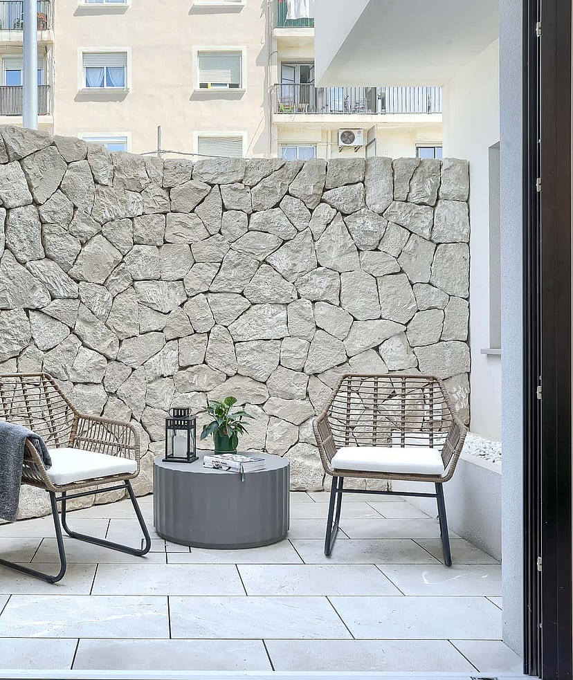 A private outdoor patio featuring a high dry-stone boundary wall, light-toned stone paving, and two rattan chairs with a small table.