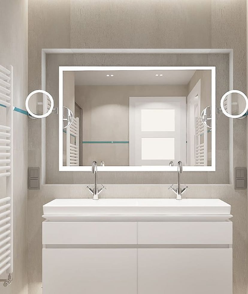 A modern bathroom featuring a white double vanity, large rectangular mirror with integrated lighting, and white heated towel rails against neutral stone-tiled walls.
