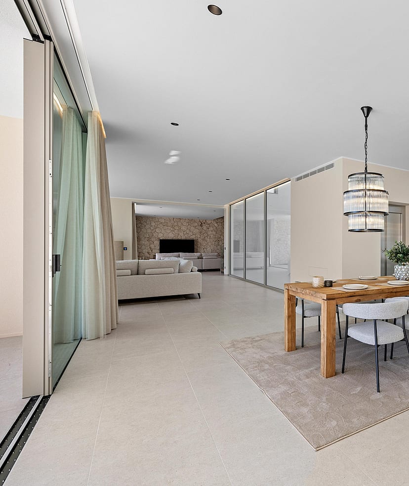 Open-plan living and dining area featuring floor-to-ceiling windows, a wooden dining table, fabric chairs, and designer lighting.