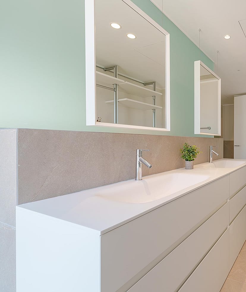 Baño moderno con doble lavabo blanco, pared verde salvia, dos espejos rectangulares y estanterías integradas.