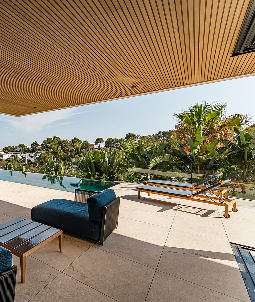 A covered terrace with a wood-slatted ceiling, limestone flooring, and blue outdoor lounge furniture overlooking a swimming pool.