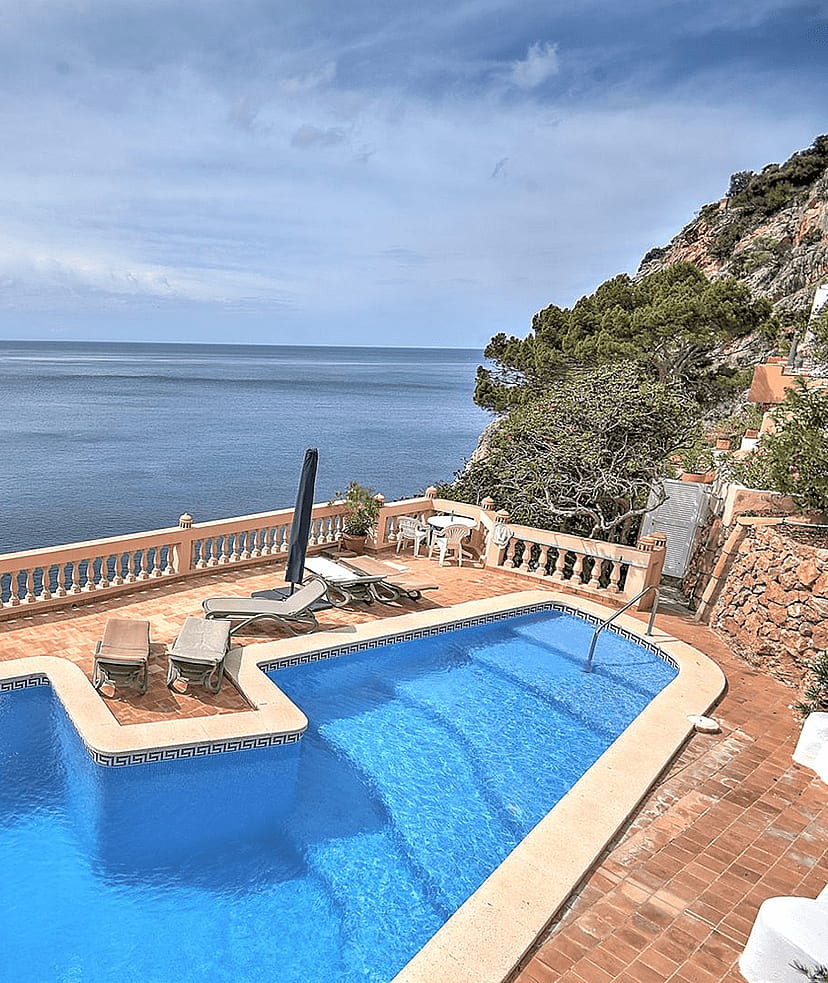 An L-shaped swimming pool with blue water sits on a terracotta-tiled terrace overlooking the sea at Cala Llamp.