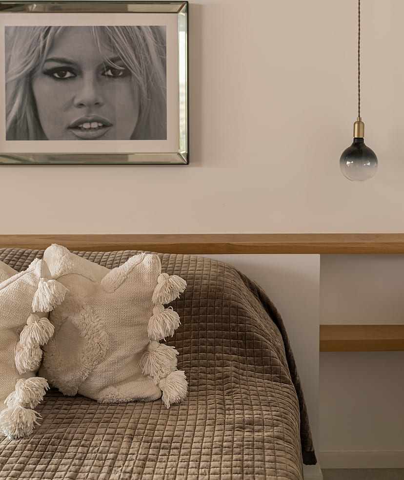 A close-up of a bedroom interior showing a textured brown bedspread, off-white decorative pillows with tassels, and a floating light-oak headboard shelf with a pendant light.