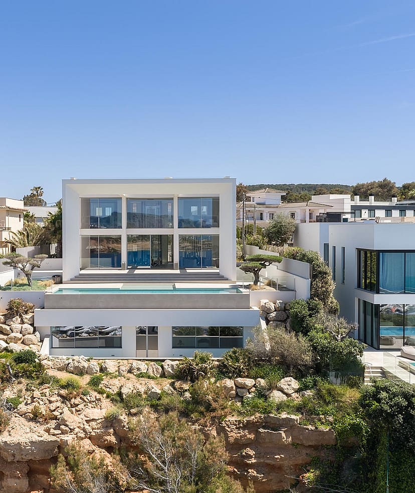 Wide exterior view of a modern white multi-level villa built into a rocky coastline with a swimming pool and glass walls.