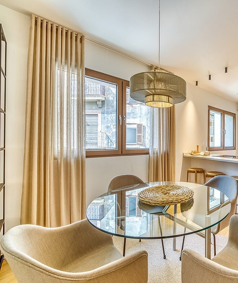 Modern open-plan kitchen with white cabinetry and dining area featuring a round glass table, beige armchairs, and floor-length curtains.