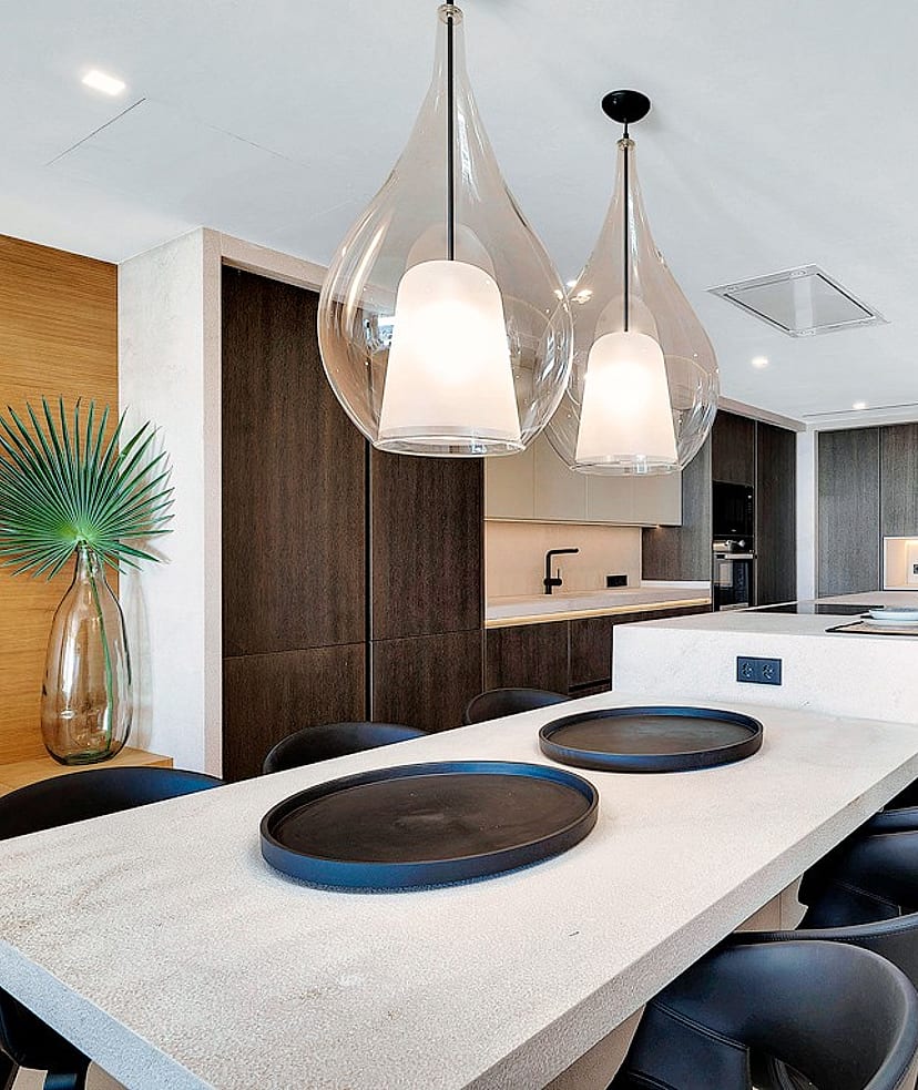 Modern open-plan kitchen featuring a long light-colored stone island, dark cabinetry, and two clear glass pendant lights.