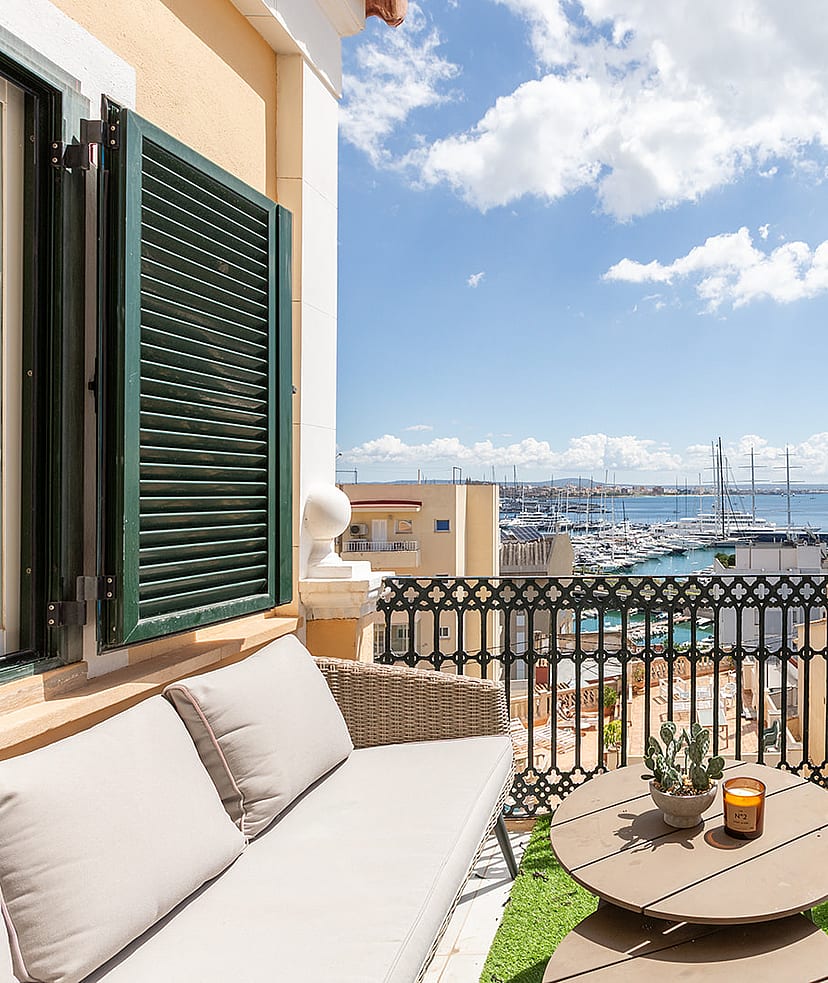 A sunny outdoor terrace featuring wicker seating, a small round table, and green shutters, overlooking a marina and sea.
