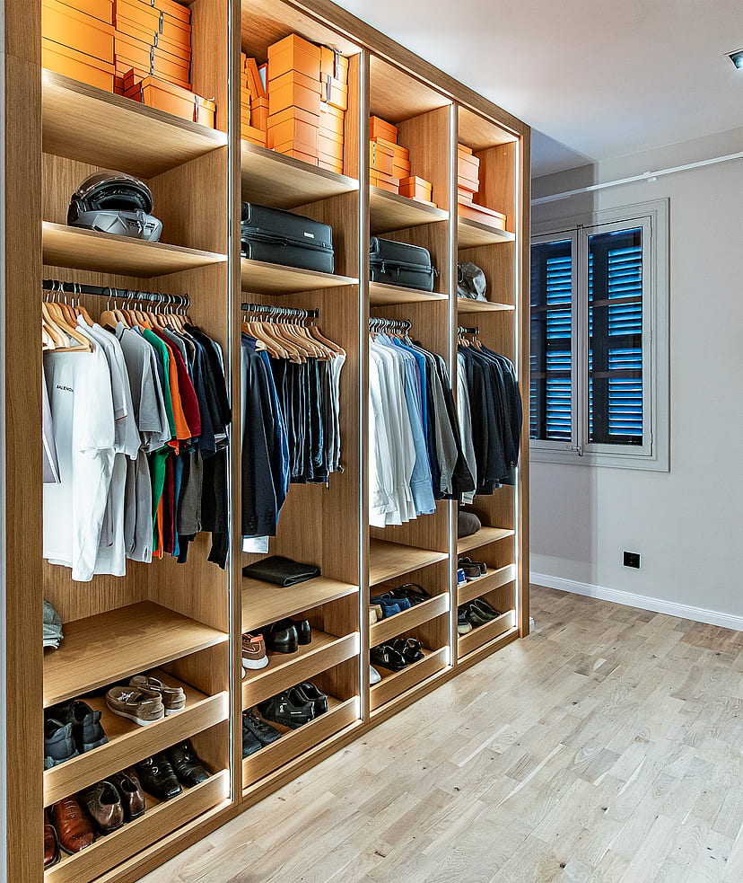 A walk-in dressing room featuring light oak cabinetry with illuminated shelving, hanging rails, and shoe racks, leading to an adjacent bedroom.