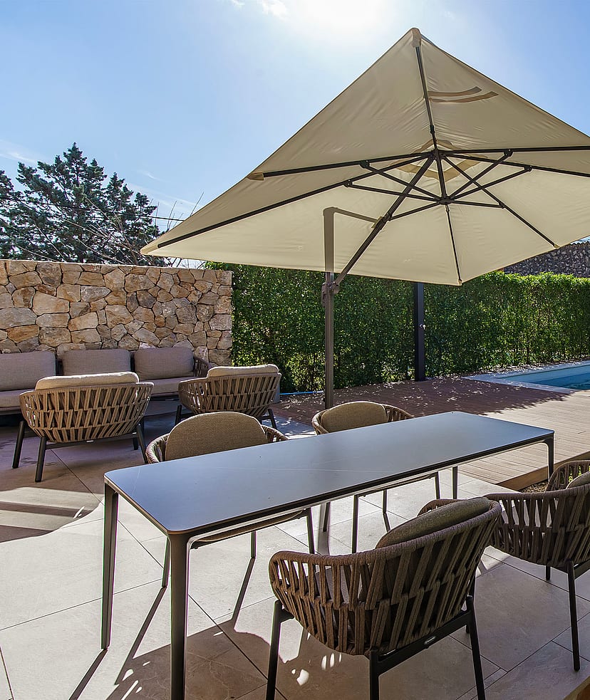 An outdoor dining terrace in Es Capdella featuring a rectangular table with woven chairs, a large beige parasol, and a small pool set against a natural stone wall.
