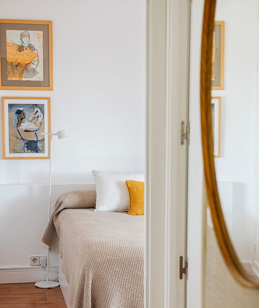 A close-up view of a bedroom interior with a bed featuring textured bedding, two framed artworks on the white wall, and a large oval mirror in the foreground.