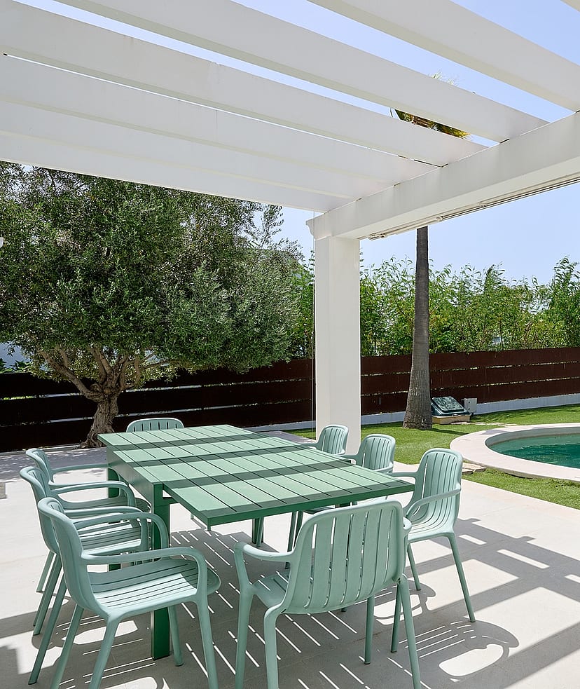Pérgola blanca sobre mesa de comedor exterior junto a piscina circular con cipreses y setos perfilados
