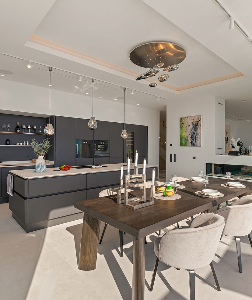 Modern open-concept interior featuring a dark grey kitchen island, a wooden dining table with light chairs, and recessed ceiling lighting.
