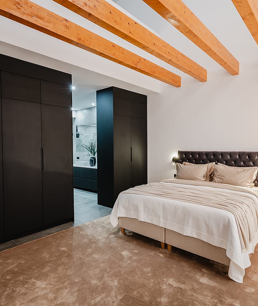 A modern bedroom with a large bed against a white wall, dark floor-to-ceiling wardrobes, and visible natural wood ceiling beams.