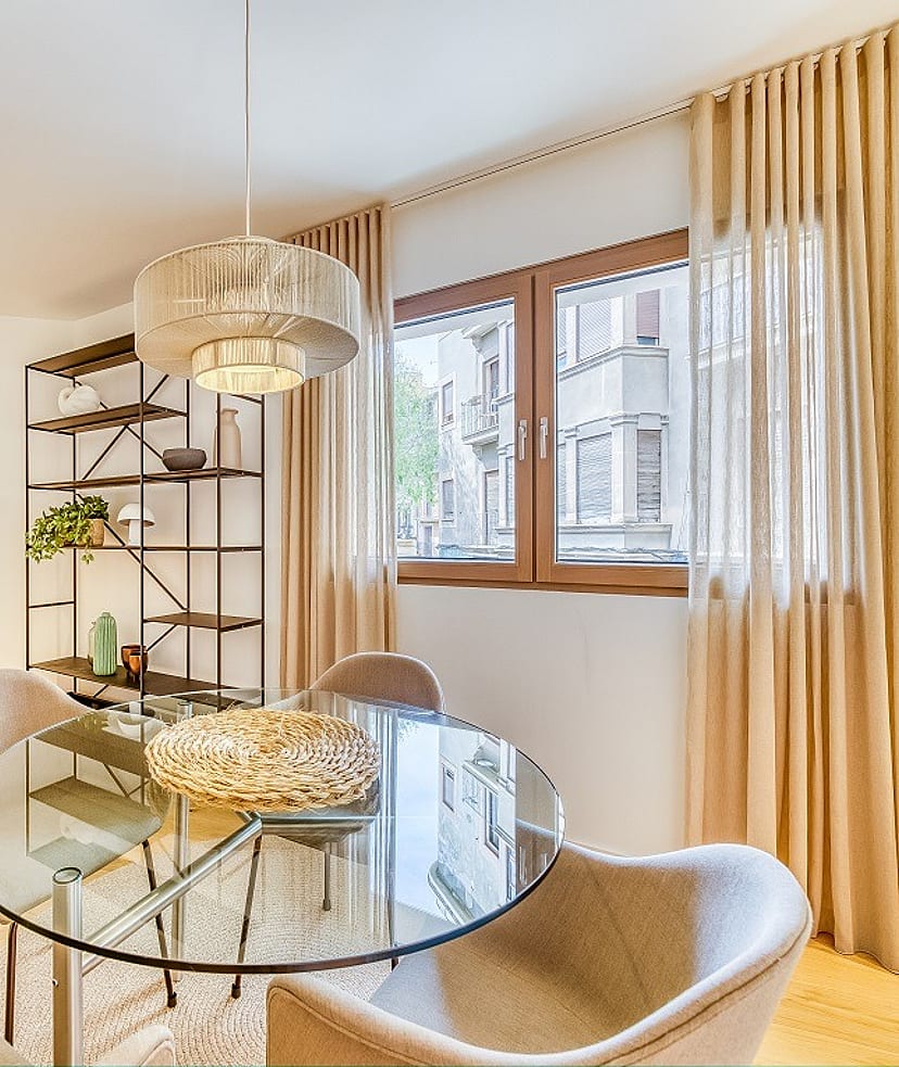 A glass dining table with four curved chairs next to a metal shelving unit in an apartment, with linen curtains framing city windows.