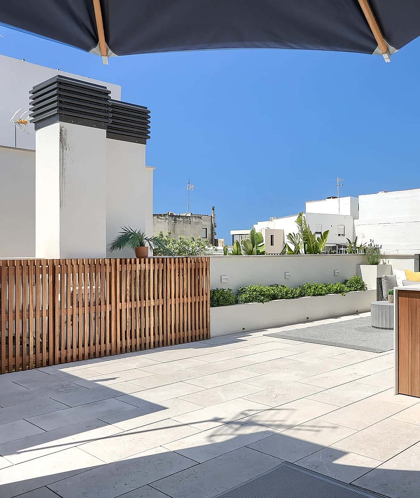 Rooftop terrace featuring a wooden outdoor kitchen cabinet with a built-in stainless steel grill, stone tile flooring, and slatted wood privacy fencing under a clear blue sky.