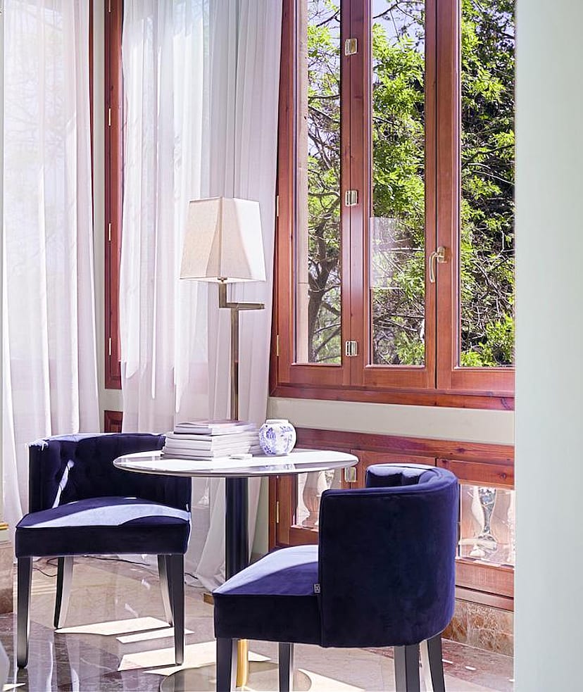 A small circular table with two blue upholstered chairs placed beside tall wood-framed windows with white curtains.