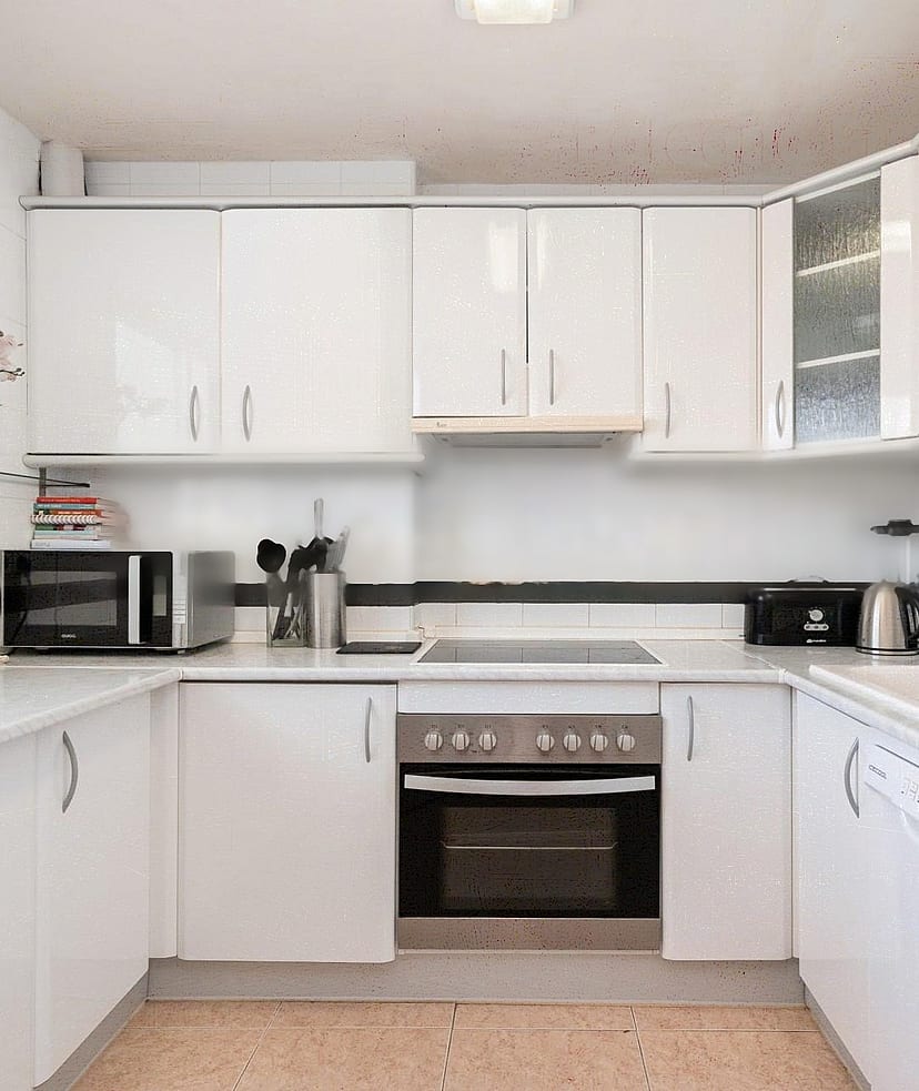 A bright galley kitchen with white cabinets, light countertops, and dark tiled backsplash, illuminated by natural light.