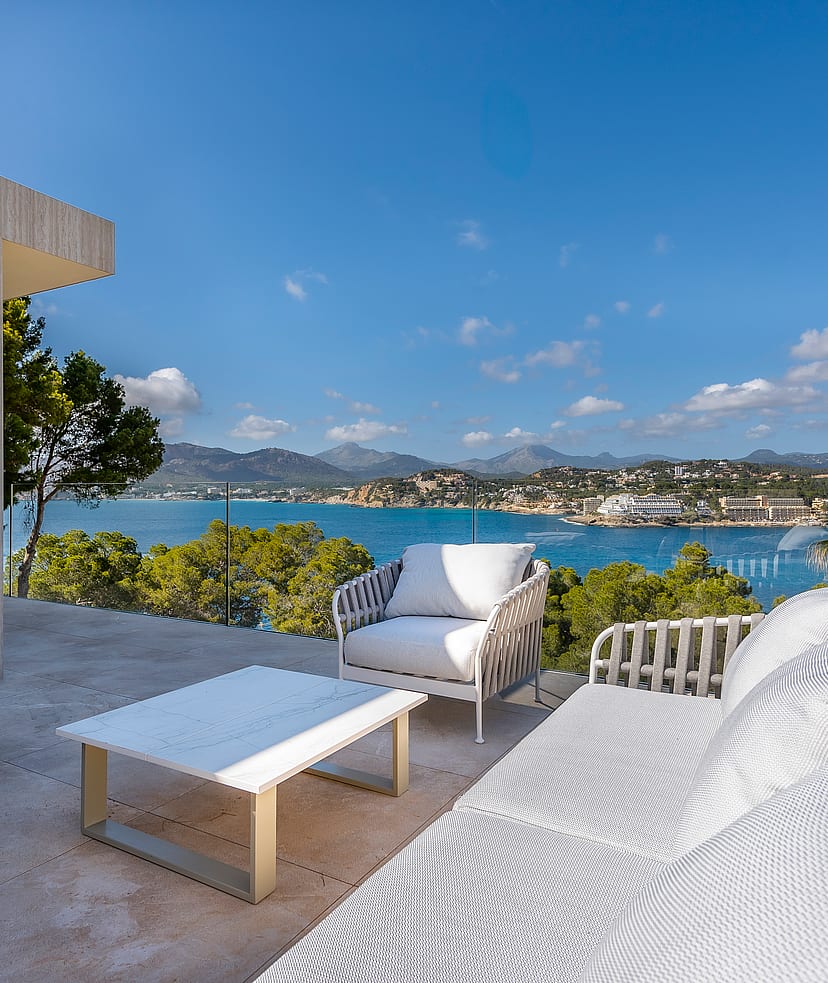 Outdoor terrace with white lounge furniture, glass railings, and a wide view of the Mediterranean bay and coastal mountains.