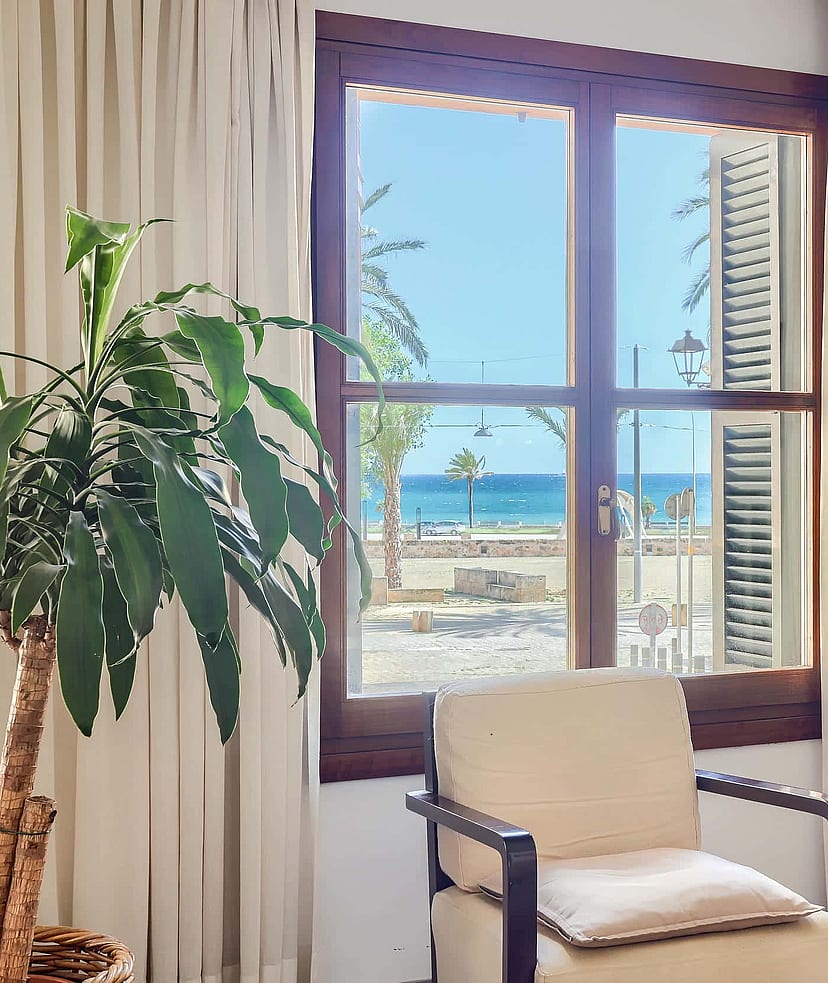 A bright living space with a wooden window frame looking out to the sea and palm trees. An armchair is placed near the window next to a houseplant and a side table with a lamp.