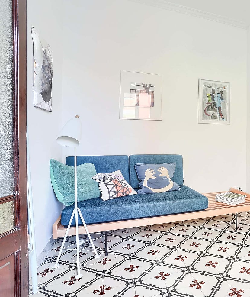 A bright living space featuring a blue sofa, a dark designer chair, and traditional patterned floor tiles.