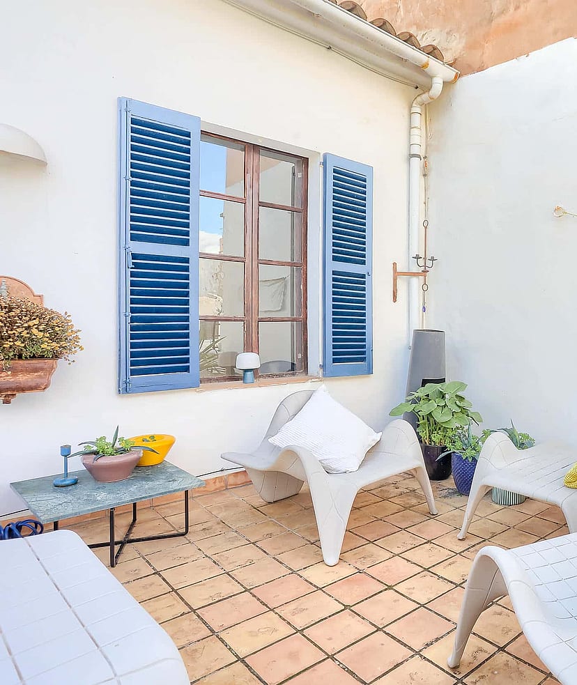 A small outdoor terrace with terracotta tiles, white plastered walls, blue wooden shutters, and modern white lounge chairs.