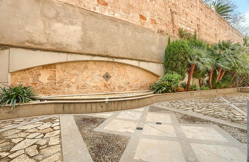 A weathered stone wall with an arched fountain feature, cobblestone patterned ground, and small green trees in a residential courtyard.