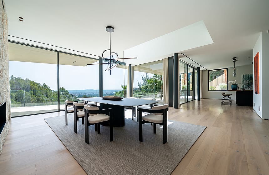 Dining room with an oval table and six chairs on a textured rug, floor-to-ceiling glass walls, and oak wood flooring.