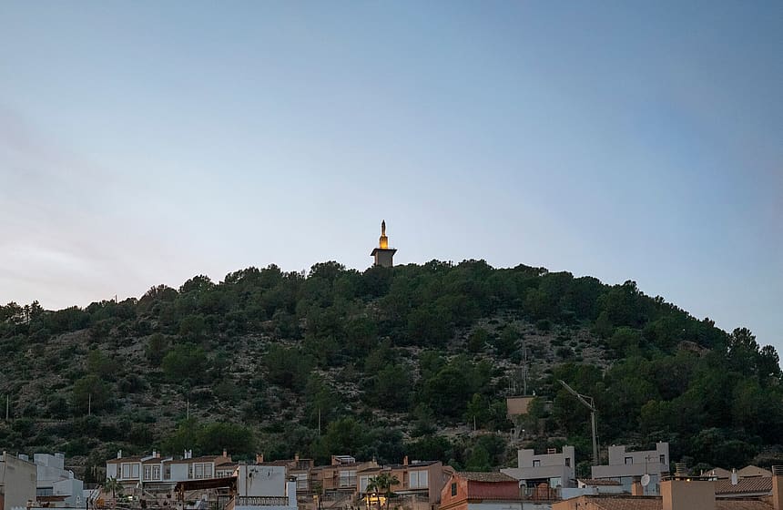 A distant view of a hillside topped with a tall landmark tower against a clear blue evening sky, seen above local rooftops.