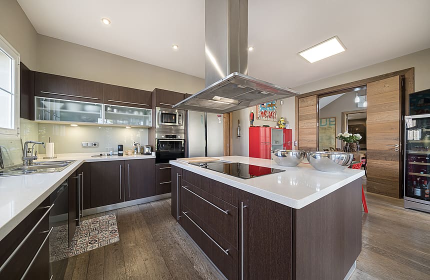 Modern kitchen featuring a large central island with white stone countertop, dark wood cabinetry, stainless steel range hood, and integrated appliances.