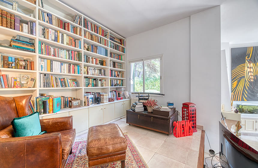 A library area with tall white bookshelves, a tan leather armchair and ottoman, a patterned rug, and a dark wooden trunk.
