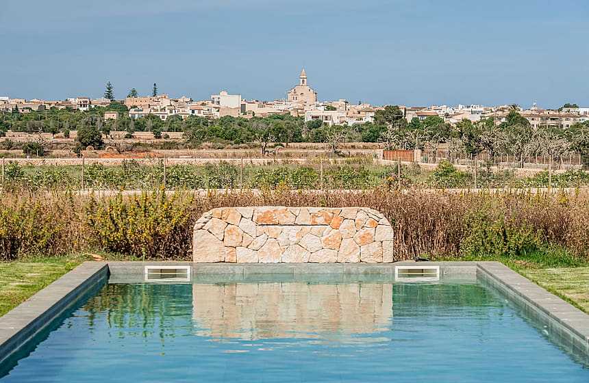 A rectangular swimming pool featuring a natural stone waterfall wall with the village of Santanyí in the background.