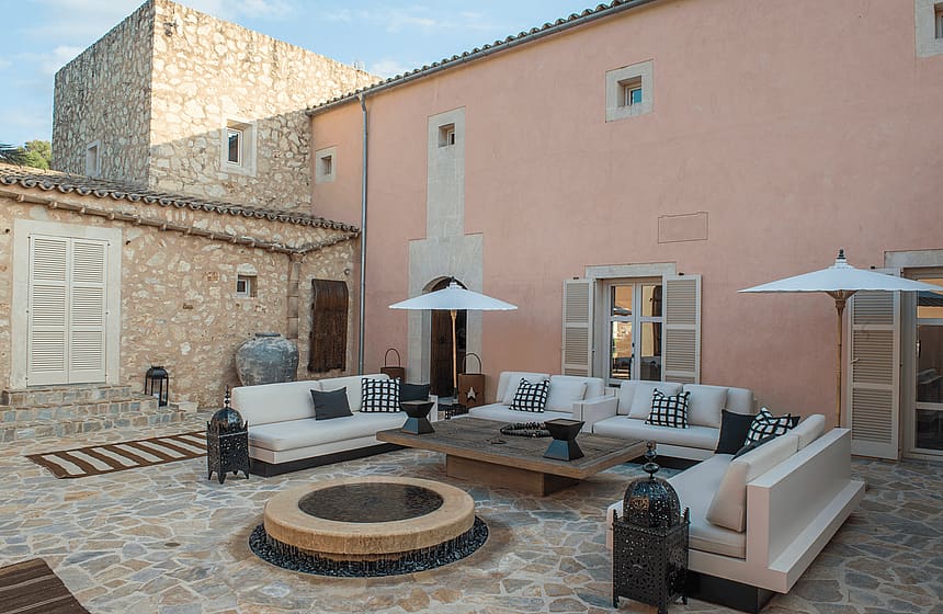 An outdoor stone courtyard of a Mallorcan finca featuring cream-colored sofas, a circular fire pit, and rustic stone walls.