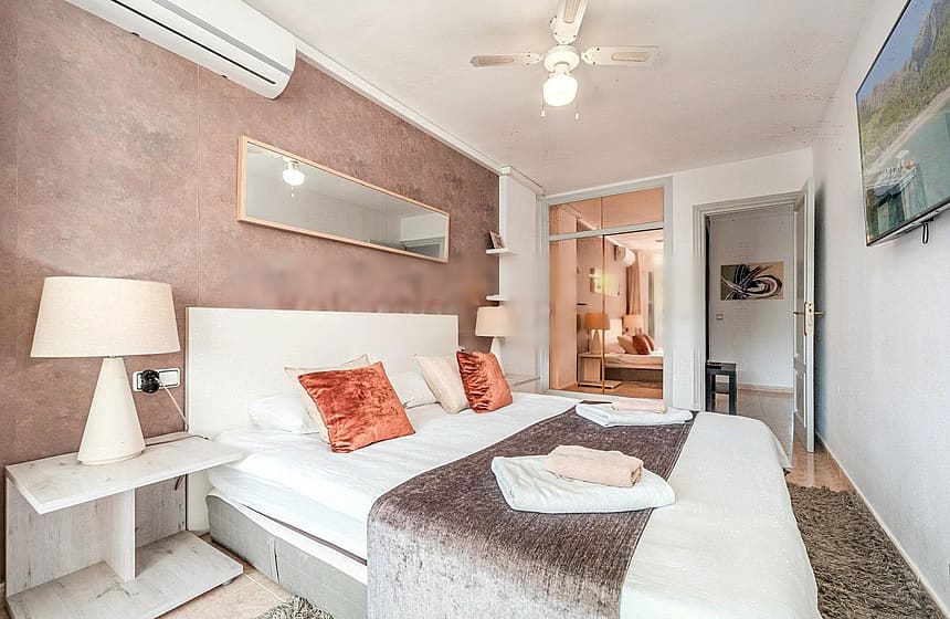 A bedroom featuring a textured dusty rose accent wall, a white bed with orange decorative pillows, a ceiling fan, and a view into a secondary room through an open doorway.