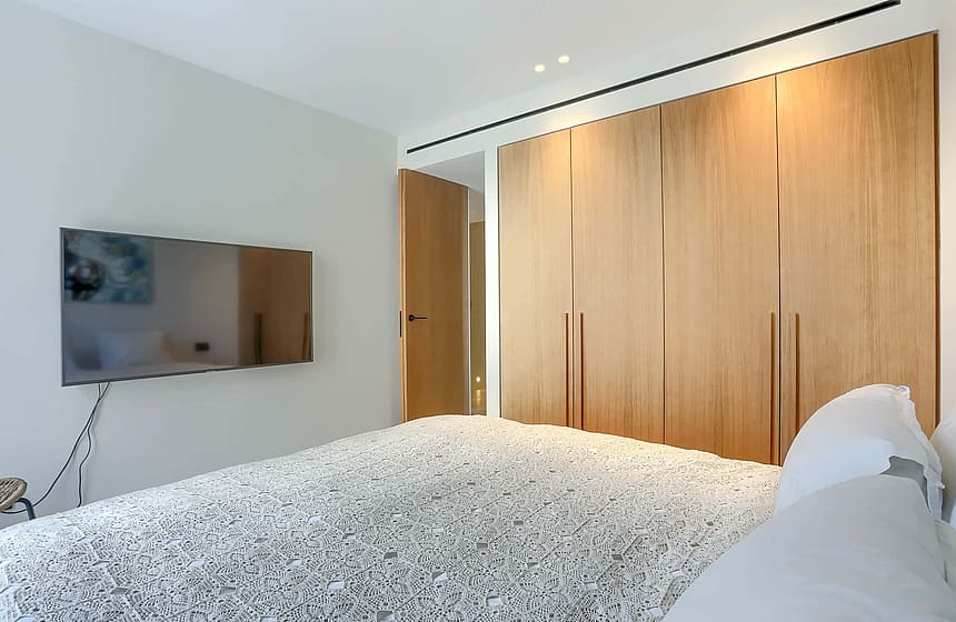 Contemporary bedroom featuring a large oak wardrobe wall, wall-mounted television, and neutral minimalist decor.