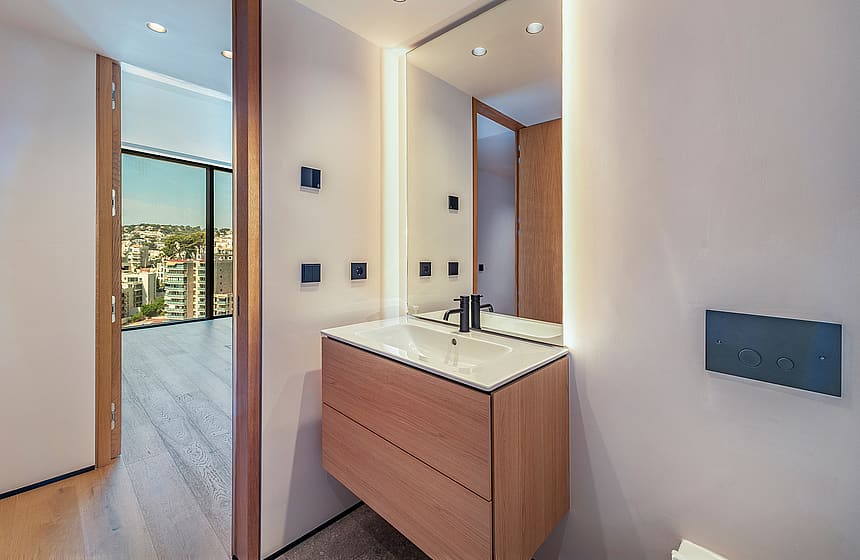 A bathroom vanity with a wooden cabinet and white countertop, featuring integrated mirror lighting and a view of the city.